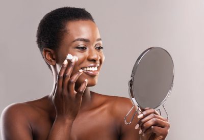 Skin care concept. Beautiful African American Woman Looking in Magnifying Mirror And Applying Face Cream Over Gray Studio Background